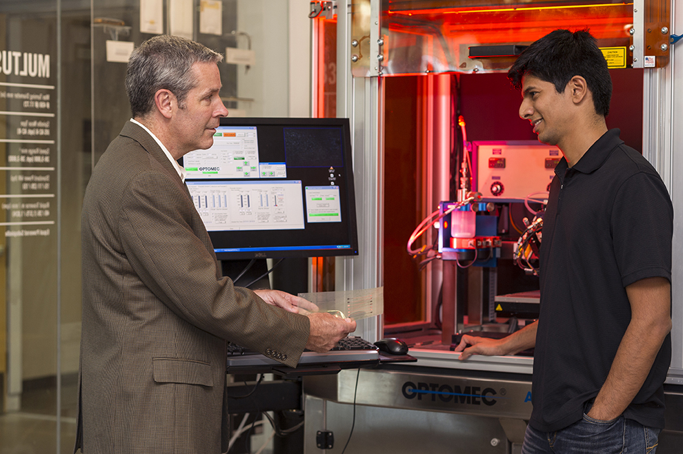 A professor talking to a student in front of an Optomec machine in the Brinkman Lab at RIT.